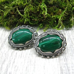 May include: A pair of silver earrings with oval-shaped malachite stones. The stones are a deep green color with visible striations. The earrings have a delicate, ornate design.