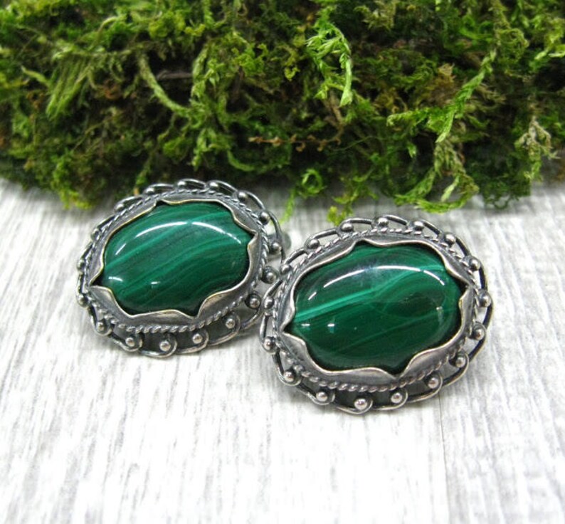 May include: A pair of silver earrings with oval-shaped malachite stones. The stones are a deep green color with visible striations. The earrings have a delicate, ornate design.