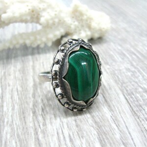 May include: A silver ring with a green malachite gemstone set in an ornate oval bezel. The ring has a delicate filigree design.