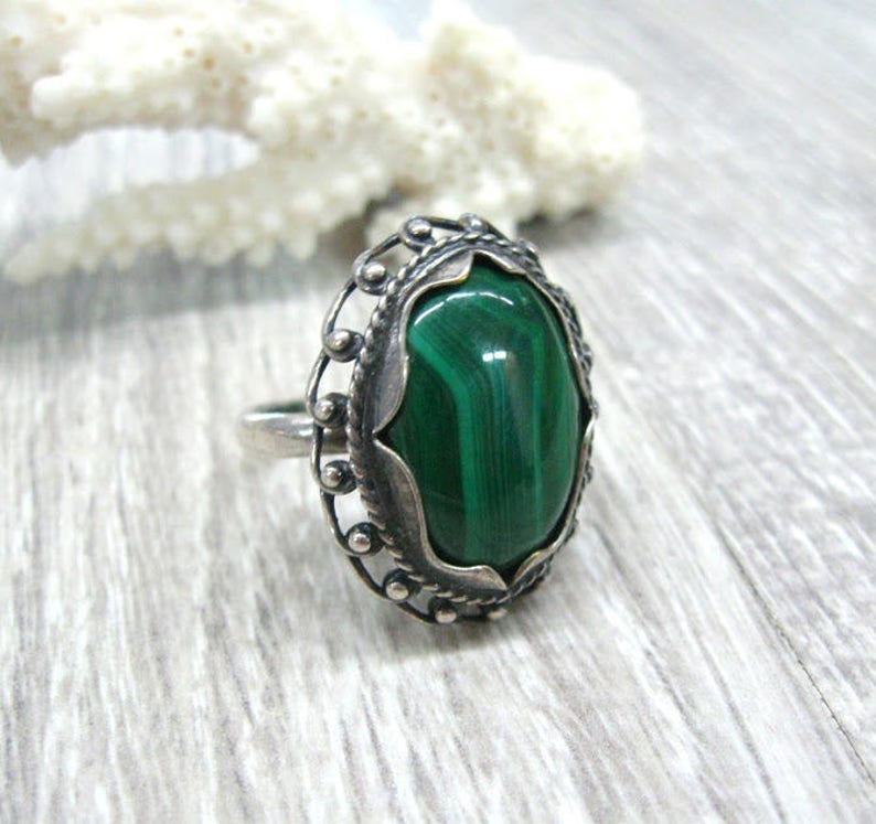 May include: A silver ring with a green malachite gemstone set in an ornate oval bezel. The ring has a delicate filigree design.