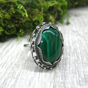 May include: A silver ring with a green malachite gemstone set in an ornate oval bezel. The ring has a delicate, filigree design.