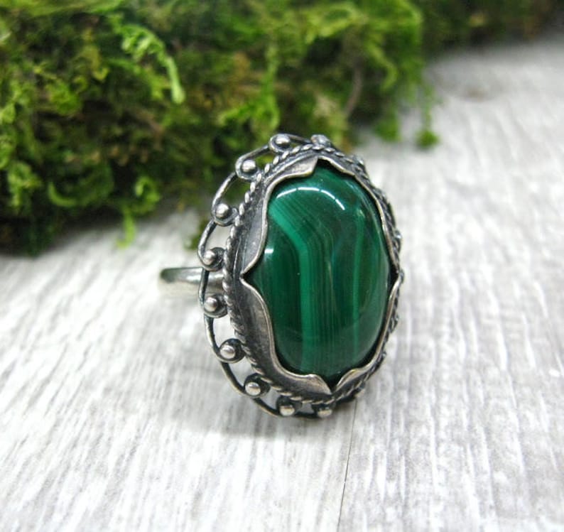 May include: A silver ring with a green malachite gemstone set in an ornate oval bezel. The ring has a delicate, filigree design.