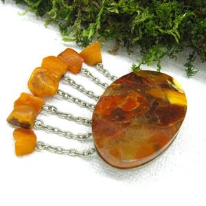 Puede incluir: Un broche vintage de &aacute;mbar con una gran piedra ovalada de &aacute;mbar y una franja de cuentas de &aacute;mbar m&aacute;s peque&ntilde;as que cuelgan de cadenas plateadas.