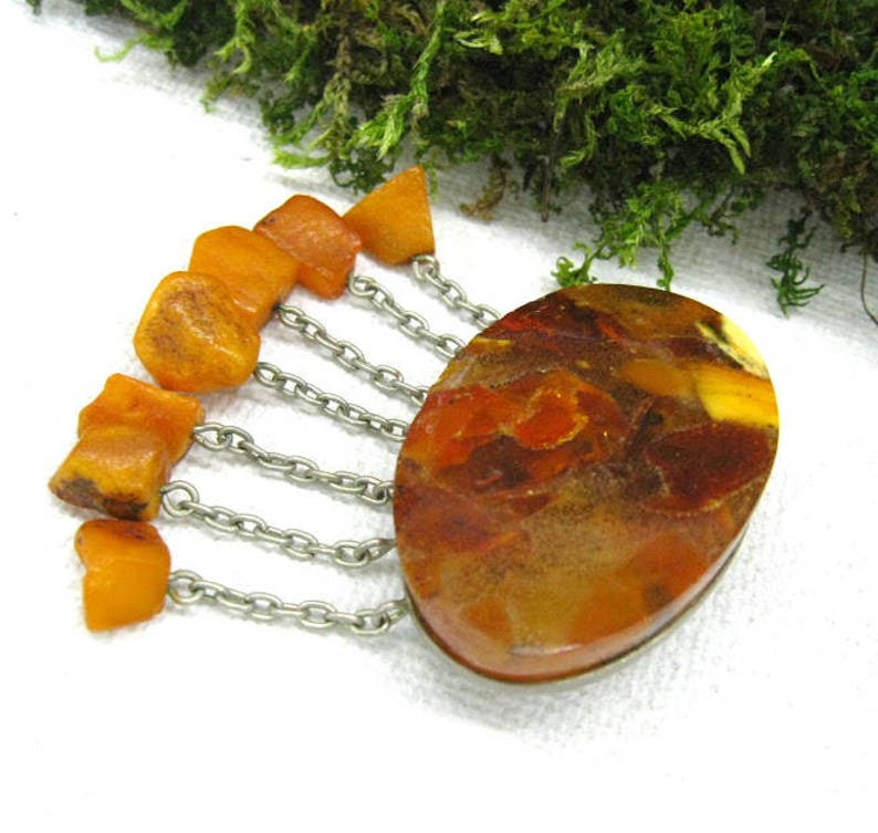 Puede incluir: Un broche vintage de &aacute;mbar con una gran piedra ovalada de &aacute;mbar y una franja de cuentas de &aacute;mbar m&aacute;s peque&ntilde;as que cuelgan de cadenas plateadas.