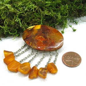 Puede incluir: Un broche grande de &aacute;mbar con una cadena de plata y cuentas de &aacute;mbar. El broche tiene forma ovalada y presenta un patr&oacute;n de m&aacute;rmol. El &aacute;mbar es de color naranja c&aacute;lido.