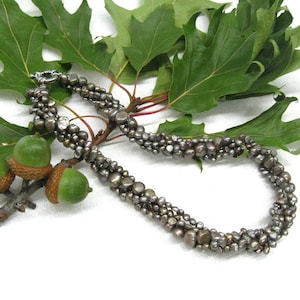 以下が含まれることがあります： 濃いグレーの淡水パールでできたマルチストランドネックレス。パールは形とサイズが不揃いで、ネックレスにユニークで自然な外観を与えています。