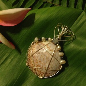 May include: A handcrafted pendant featuring a seashell wrapped in silver wire. The shell is adorned with pearls and small black beads, and a silver star charm. The pendant rests on a large green leaf, with a pink and white flower in the background.