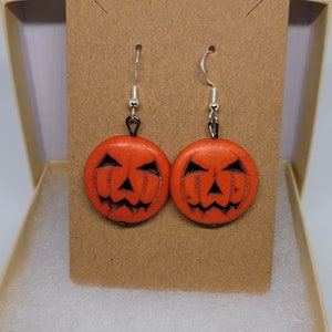 May include: A pair of orange pumpkin-shaped earrings with black painted jack-o'-lantern faces. The earrings are on a brown card in a white gift box.