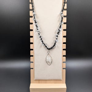 May include: A necklace featuring a black cord knotted with small, clear, irregular crystal chips along its length. The necklace includes a central pendant shaped like an elongated oval, constructed from silver-toned wire containing a cluster of clear crystal chips.