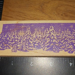 Könnte beinhalten: Ein Holzstempel mit einem violetten Hintergrund und einem Design eines schneebedeckten Waldes mit Sternen.