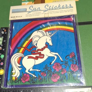 Puede incluir: Un paquete de "Sun Stickers" con un unicornio blanco con una cinta roja, un arcoíris y rosas rosas. El paquete dice "Made in U.S.A." y "¡Una calcomanía transparente que es removible y reutilizable! Decora tus ventanas con y aplica a la ventana."
