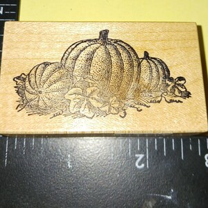 May include: A wooden stamp featuring a detailed illustration of three pumpkins and leaves. The pumpkins are of varying sizes, with the largest in the center. The stamp is rectangular and made of light-colored wood.
