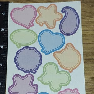 Puede incluir: Una hoja de pegatinas de colores en varias formas, incluyendo corazones, estrellas y bocadillos. Cada pegatina tiene un contorno de color y un interior de color pastel. Las pegatinas están sobre un fondo blanco.
