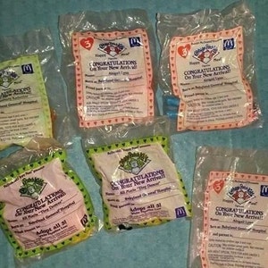 May include: A collection of six sealed Happy Meal toys from McDonald's, featuring Cabbage Patch Kids. Each package has a congratulatory message for a new arrival, with names and birth locations listed. The packages are in various colors, including pink, green, and yellow.