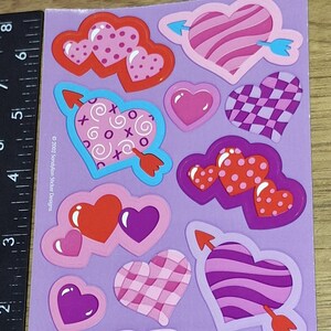 Puede incluir: Hoja de pegatinas con forma de corazón en varios diseños y colores. Incluye corazones con rayas, lunares y flechas. Las pegatinas son en tonos de rosa, rojo y morado, con acentos azules. La hoja está sobre un fondo morado.
