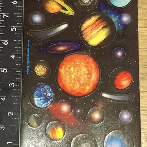 Puede incluir: Hoja de pegatinas de planetas coloridos, incluyendo Saturno, el sol y varias galaxias. Las pegatinas están sobre un fondo negro con una regla en el lateral. El texto "©2001 Sandylion Sticker Designs" es visible. Medidas en centímetros.