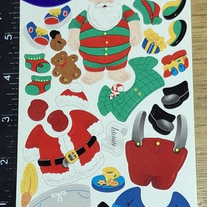 Puede incluir: Una hoja de pegatinas titulada "Dress-A-Santa" con un Papá Noel de dibujos animados y varias pegatinas de ropa y accesorios. Incluye un Papá Noel a rayas rojas y verdes, abrigo rojo, botas negras y un hombre de jengibre. ©2001 Sandylion Sticker Designs.