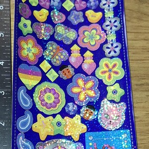 Puede incluir: Una hoja de pegatinas coloridas y brillantes sobre un fondo azul. Las pegatinas presentan varios diseños, incluyendo flores, mariposas, estrellas y una cara de gato. El texto "© 2001 Sandylion Sticker Designs" es visible.