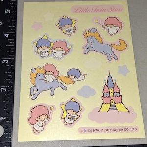 Könnte beinhalten: Ein Bogen mit Little Twin Stars Aufklebern auf gelbem Hintergrund. Die Aufkleber zeigen Zeichentrickfiguren, Einhörner, Sterne und ein Schloss. Die Figuren haben rosa und blaue Haare. Der Text "Little Twin Stars" steht oben. Copyright 1976, 1986 Sanrio Co., Ltd.