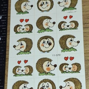 Puede incluir: Hoja de pegatinas con erizos de dibujos animados. Los erizos tienen púas marrones, caras blancas y ojos negros. Algunas pegatinas muestran erizos con corazones. El texto "11-370" está impreso en la hoja.