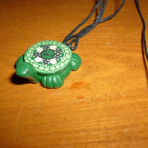Op de afbeelding: Een groene schildpad hanger ketting. De schildpad hanger heeft een patroon schild met witte en zwarte bloemmotieven. Het lichaam van de schildpad is effen groen. De ketting heeft een zwart koord.