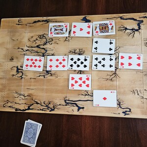 May include: A wooden board game with playing cards arranged on it. The board has a light brown finish with dark, lightning-like patterns. Playing cards are laid out, showing various suits and numbers, including hearts, diamonds, clubs, and spades.