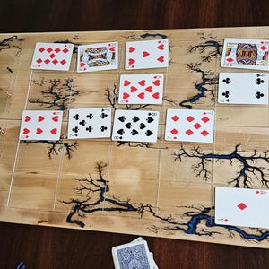 May include: A wooden game board with a grid pattern, featuring playing cards arranged on its surface. The board has a light wood grain with dark, lightning-like patterns. Several playing cards are scattered around the board.