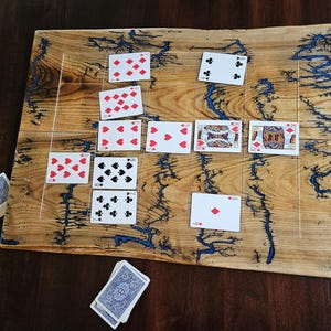 May include: A wooden board game with a natural wood grain finish and blue resin accents. Playing cards are arranged on the board, with additional cards stacked nearby. The game appears to be a variation of a card game.