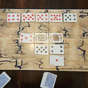 May include: A wooden game board with a light wood grain finish, featuring a decorative tree-like design with dark blue accents. Playing cards are arranged on the board, with additional cards nearby. The game board is designed for card games.