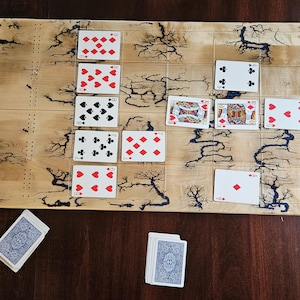 May include: A wooden cribbage board with a unique lightning pattern, featuring playing cards arranged on the board. Several decks of cards are also visible. The board is set on a dark wood surface.