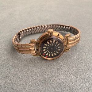 May include: A gold-toned wristwatch with a black face and a gold-toned metal band. The watch face has a unique design with a black and white pattern and a red heart.