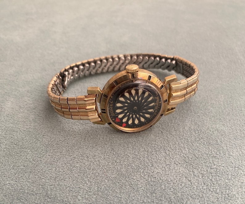 May include: A gold-toned wristwatch with a black face and a gold-toned metal band. The watch face has a unique design with a black and white pattern and a red heart.