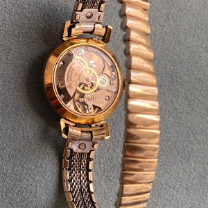 May include: A gold-toned wristwatch with a round face and a metal band. The watch face is clear and shows the inner workings of the watch. The band is a combination of a mesh design and a more traditional link design.