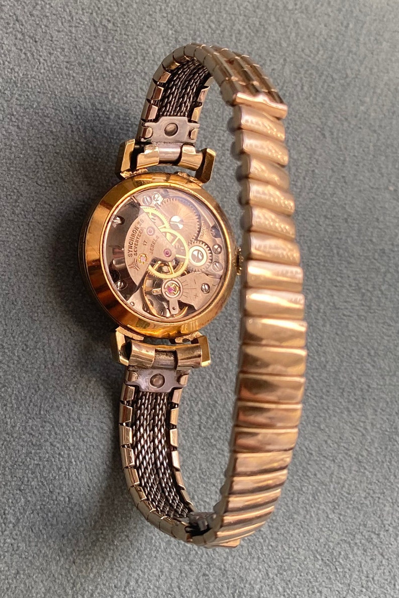 May include: A gold-toned wristwatch with a round face and a metal band. The watch face is clear and shows the inner workings of the watch. The band is a combination of a mesh design and a more traditional link design.
