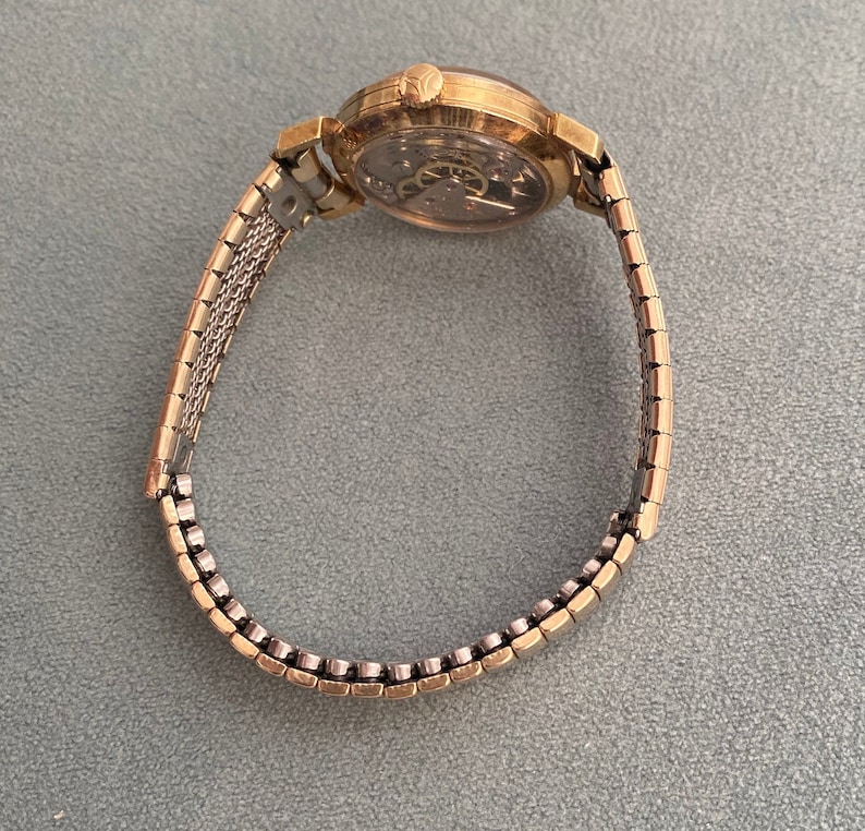 May include: A gold-toned wristwatch with a round face and a metal band. The watch face has a white background with black markings and hands.