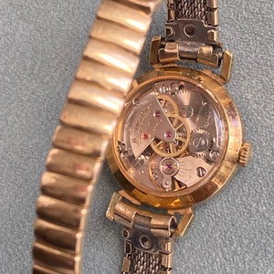 May include: A gold-toned wristwatch with a round face and a metal band. The back of the watch is open, revealing the inner workings of the watch.
