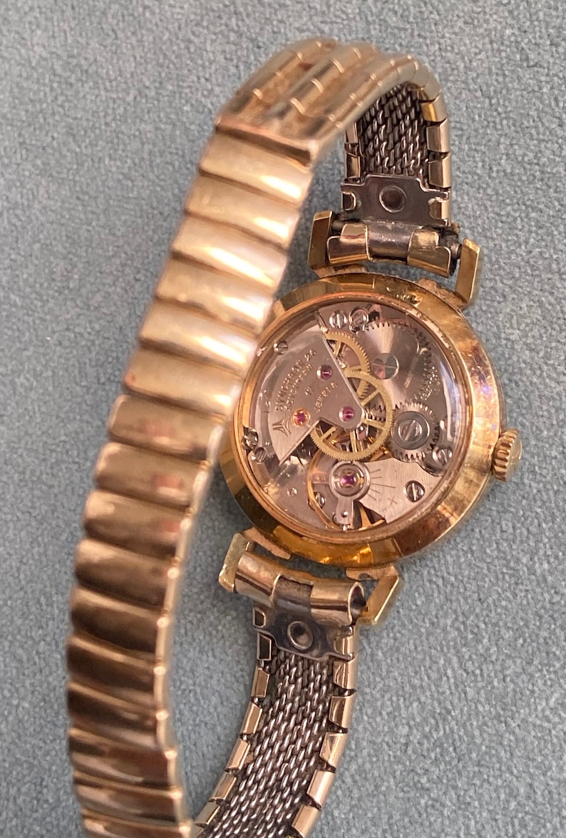 May include: A gold-toned wristwatch with a round face and a metal band. The back of the watch is open, revealing the inner workings of the watch.