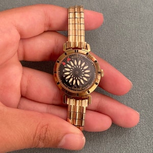 May include: A gold-toned wristwatch with a black face and a white and gold design. The watch face says "Borel Cocktail" and has a red heart on the dial.