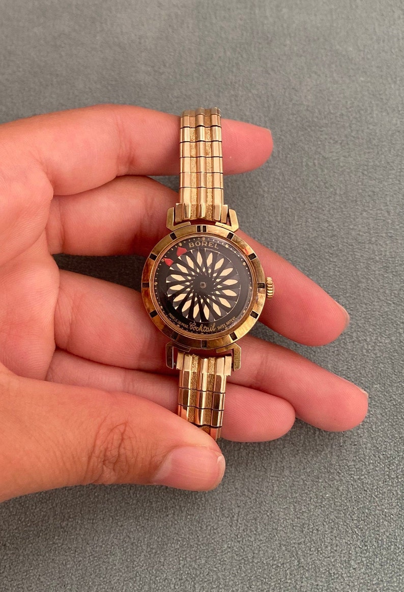 May include: A gold-toned wristwatch with a black face and a white and gold design. The watch face says "Borel Cocktail" and has a red heart on the dial.