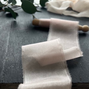 May include: A pale pink silk ribbon with a subtle shimmer, rolled on a wooden dowel. The ribbon is on a black slate surface.