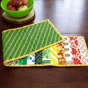 Peut inclure: Un chemin de table en tissu matelassé avec une grande section verte à rayures diagonales sombres et une bordure jaune. La partie restante présente une série de bandes de tissu verticales aux motifs variés, incluant des designs floraux, cachemire et géométriques.