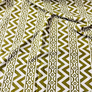 May include: Close-up of white fabric featuring a repeating pattern of olive green zigzags and vertical stripes of circles and half-circles. The fabric is folded, creating shadows and texture.