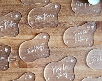 May include: A set of 15 clear acrylic place cards with white star accents and names written in white. The place cards are shaped like irregular, organic shapes.