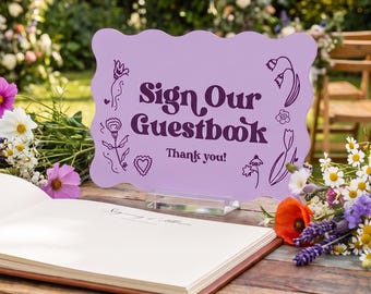 Könnte beinhalten: Ein hellviolettes, rechteckiges Schild mit gewelltem Rand, das den dunkelvioletten Text "Sign Our Guestbook" und "Thank you!" sowie florale Strichzeichnungen zeigt. Das Schild steht auf einem Holztisch neben einem offenen Gästebuch.
