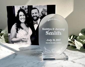 Könnte beinhalten: Eine ovale Auszeichnung aus weißem Acryl mit dem Text "Joey's Sister", "Kaitlyn & Jeremy Smith", "July 31, 2017" und "Palm Beach, Aruba", der darauf graviert ist. Die Auszeichnung steht auf einem klaren Acrylsockel.