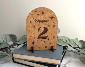 May include: An arched wooden sign featuring the engraved text Chapter 2, surrounded by various star-shaped patterns. The sign is supported by two small dark brown wooden stands, creating a display piece with a natural wood grain finish.