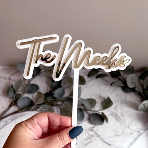 May include: White acrylic cake topper with the words "The Meeks" in gold mirror acrylic.