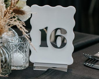 May include: A white scalloped table number sign featuring the black number 16, displayed on a clear acrylic base. Next to it is a small glass vase containing white roses and dried grasses, alongside a clear textured glass votive candle holder.