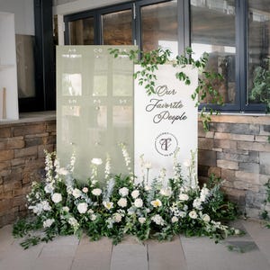 K&ouml;nnte beinhalten: Ein Hochzeits-Sitzplan mit hellgr&uuml;nem Hintergrund und transparenten Tafeln f&uuml;r die Namen der G&auml;ste. Das Schild ist mit wei&szlig;en Blumen und Gr&uuml;n geschm&uuml;ckt. Ein zweites Schild lautet "Our Favorite People."