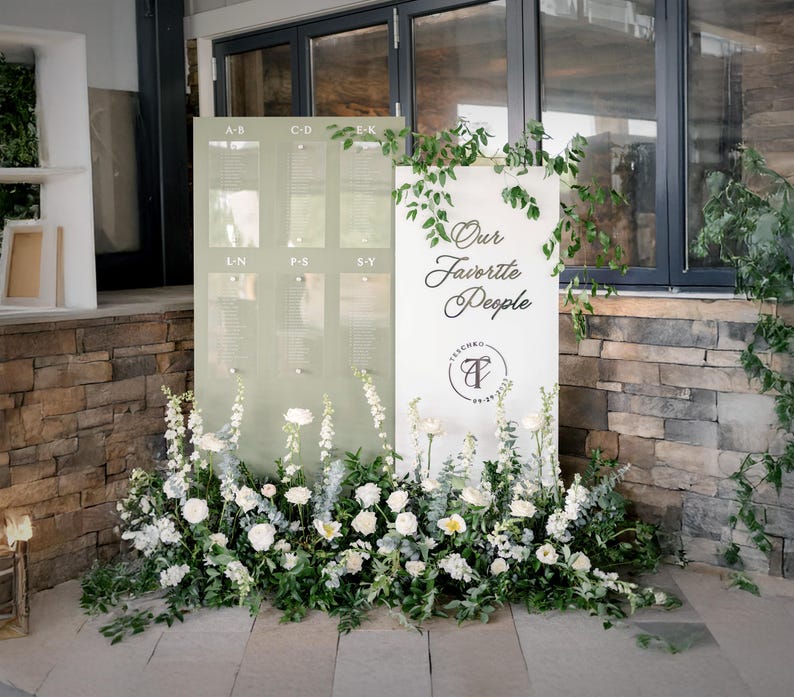 K&ouml;nnte beinhalten: Ein Hochzeits-Sitzplan mit hellgr&uuml;nem Hintergrund und transparenten Tafeln f&uuml;r die Namen der G&auml;ste. Das Schild ist mit wei&szlig;en Blumen und Gr&uuml;n geschm&uuml;ckt. Ein zweites Schild lautet "Our Favorite People."
